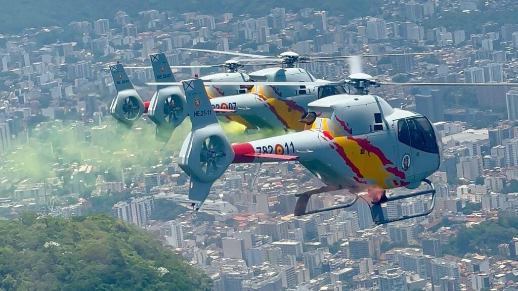 Helicópteros de la Patrulla ASPA en vuelo de formación cerrado lanzando humo de colores sobre una ciudad.
