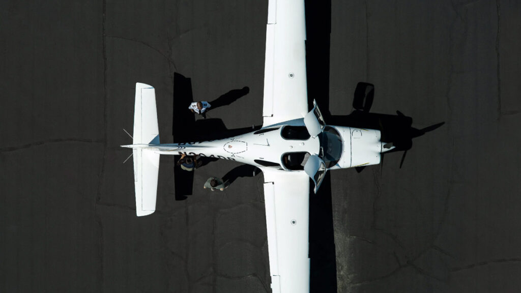 Vista aérea cenital del nuevo avión de entrenamiento Cirrus TRAC10 estacionado en plataforma con las puertas de cabina abiertas y tres personas a su alrededor.