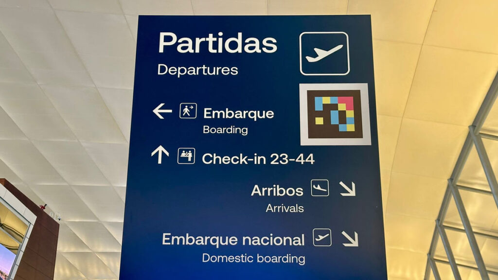 Cartel indicador direccional azul en el Aeropuerto de Carrasco con un código de accesibilidad NaviLens.