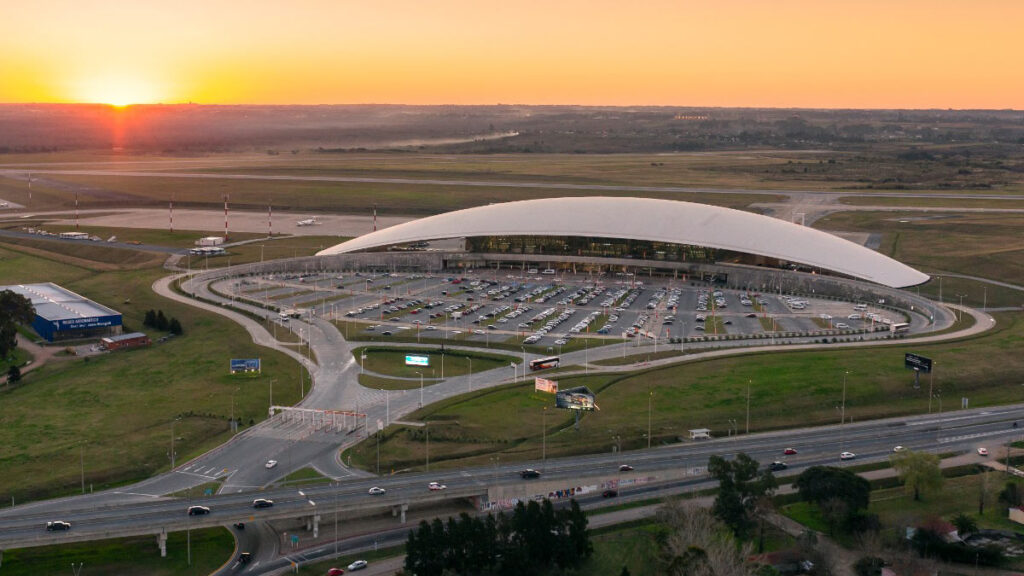 Vista aérea de la terminal del Aeropuerto Internacional de Carrasco, mostrando su techo curvo, el estacionamiento y los accesos vehiculares durante el atardecer.