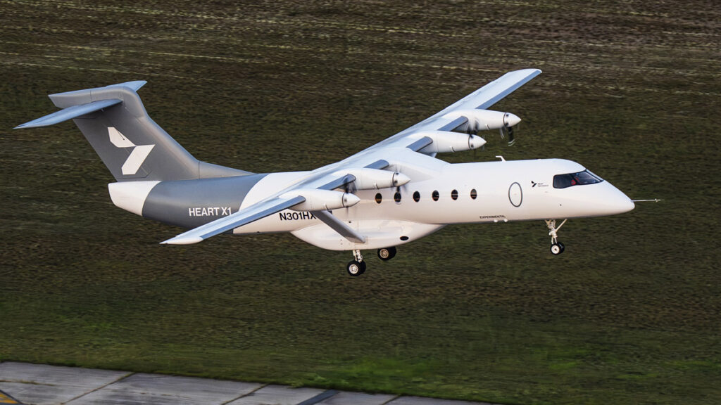 Avión eléctrico Heart X1 en vuelo a baja altitud con el tren de aterrizaje desplegado sobre un terreno de césped y el borde de una pista