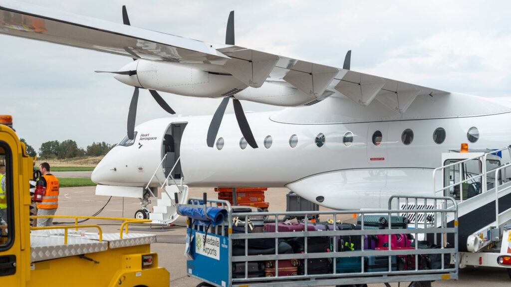 Avión eléctrico de Heart Aerospace estacionado en plataforma con la puerta de pasajeros abierta, rodeado de equipos de apoyo terrestre y carros de equipaje