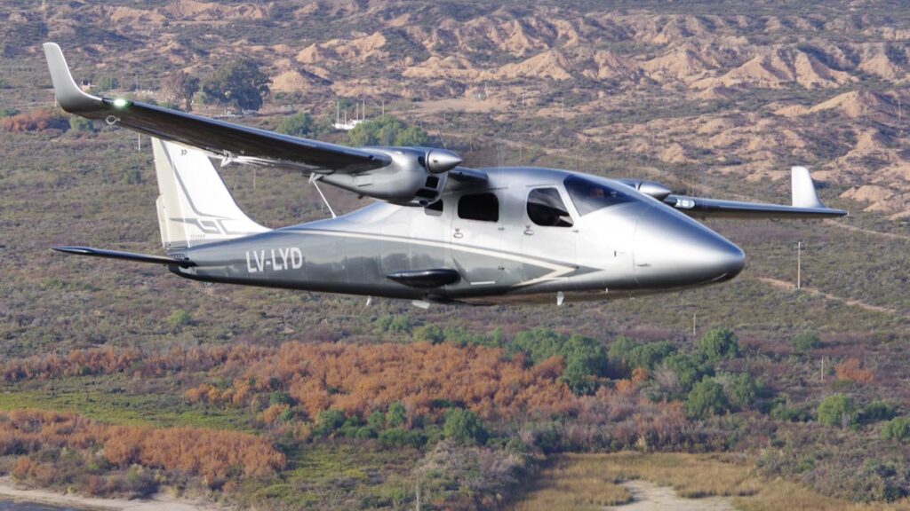 Avión bimotor Tecnam P2006T NG SPORT con matrícula LV-LYD en pleno vuelo sobre un paisaje montañoso.
